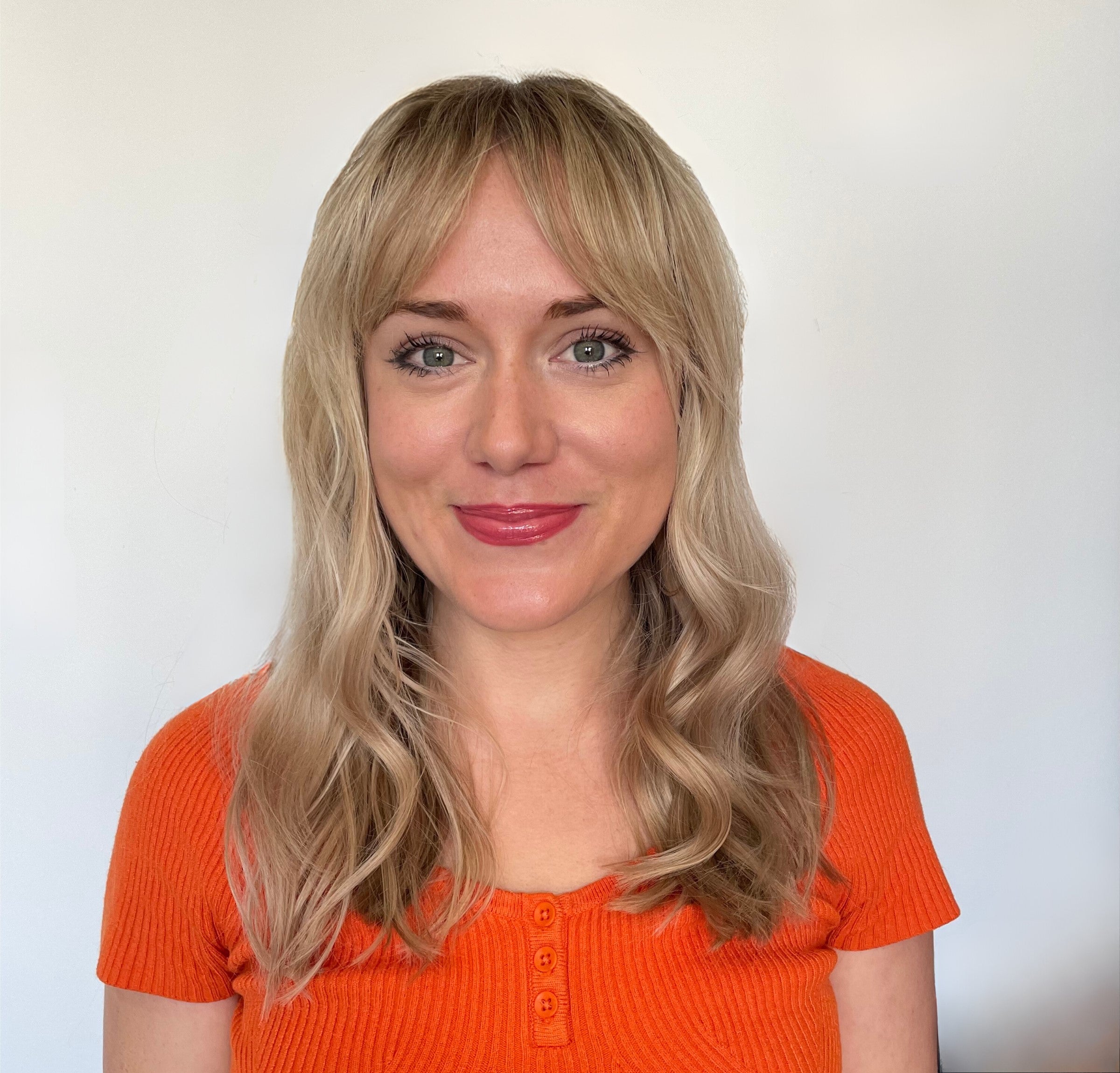 A female presenting individual with blond hair, looking into the camera and smiling. She is wearing an orange shirt.