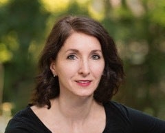 A female presenting individual with shoulder length brown hair smiling into the camera. Sitting in front of greenry.