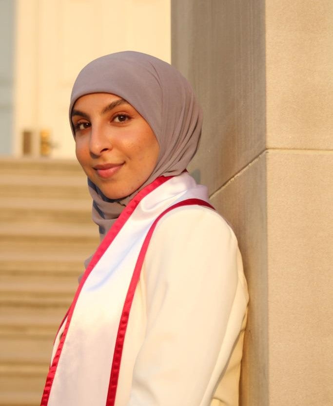 A female presenting individual standing in front of steps. Wearing a headdress. they are smiling into the camera.