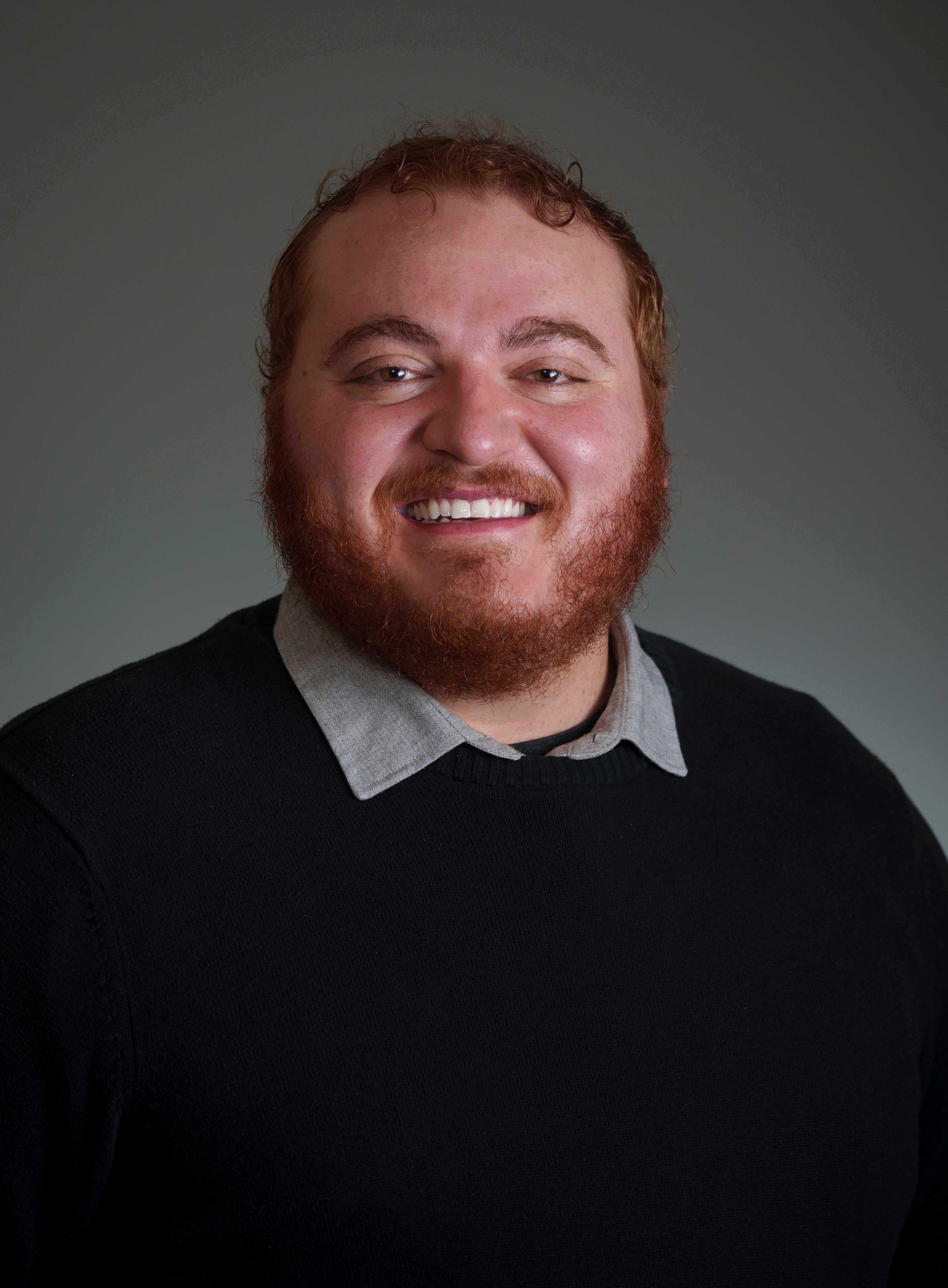 A male presenting individual, wearing a long sleeve black sweater. He has red hair and beard. He is smiling into the camera. 