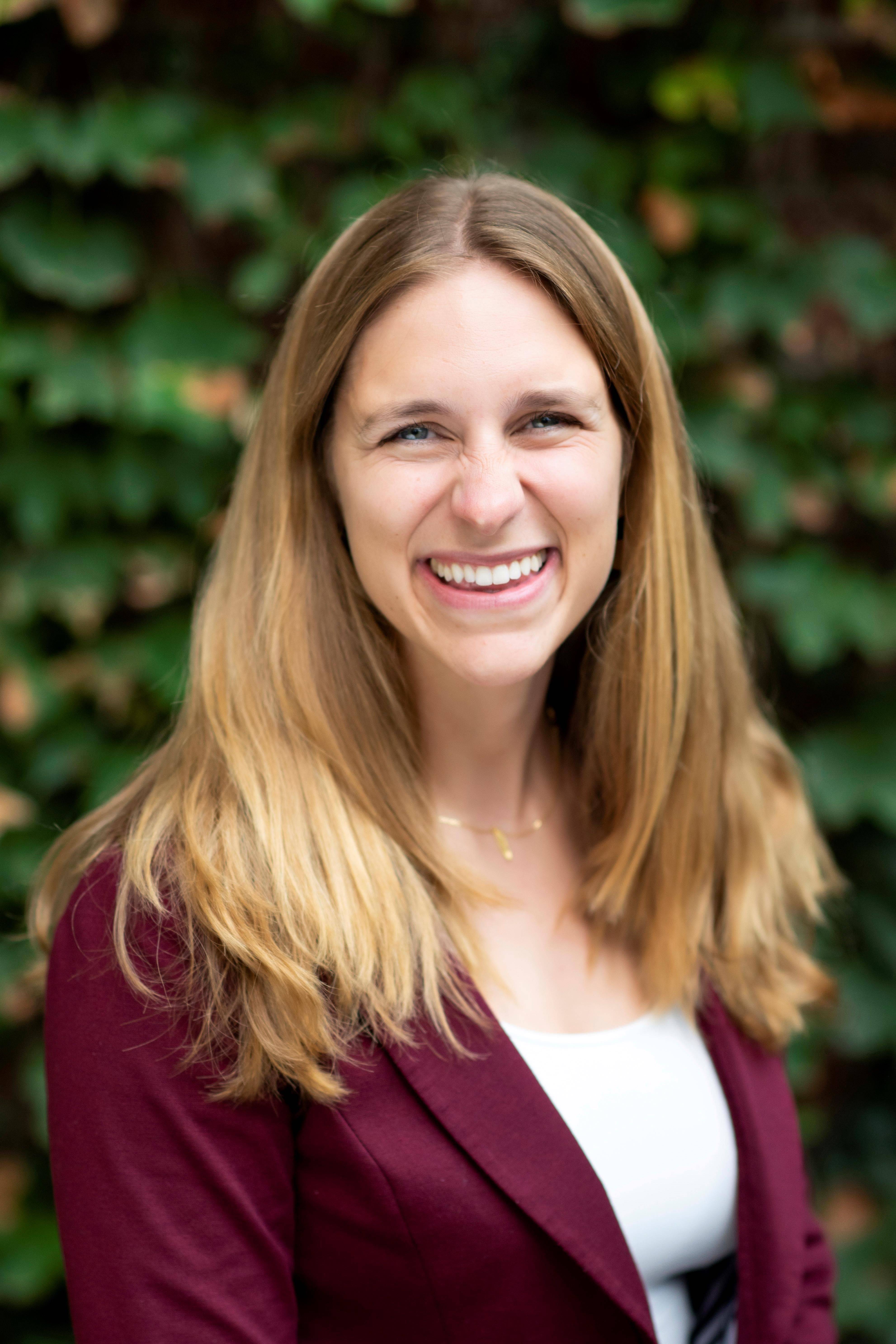 A female presenting individual with shoulder length dark blond hair is wearing a burgundy blazer and standing in front of a shrub. She is smiling into the camera. 