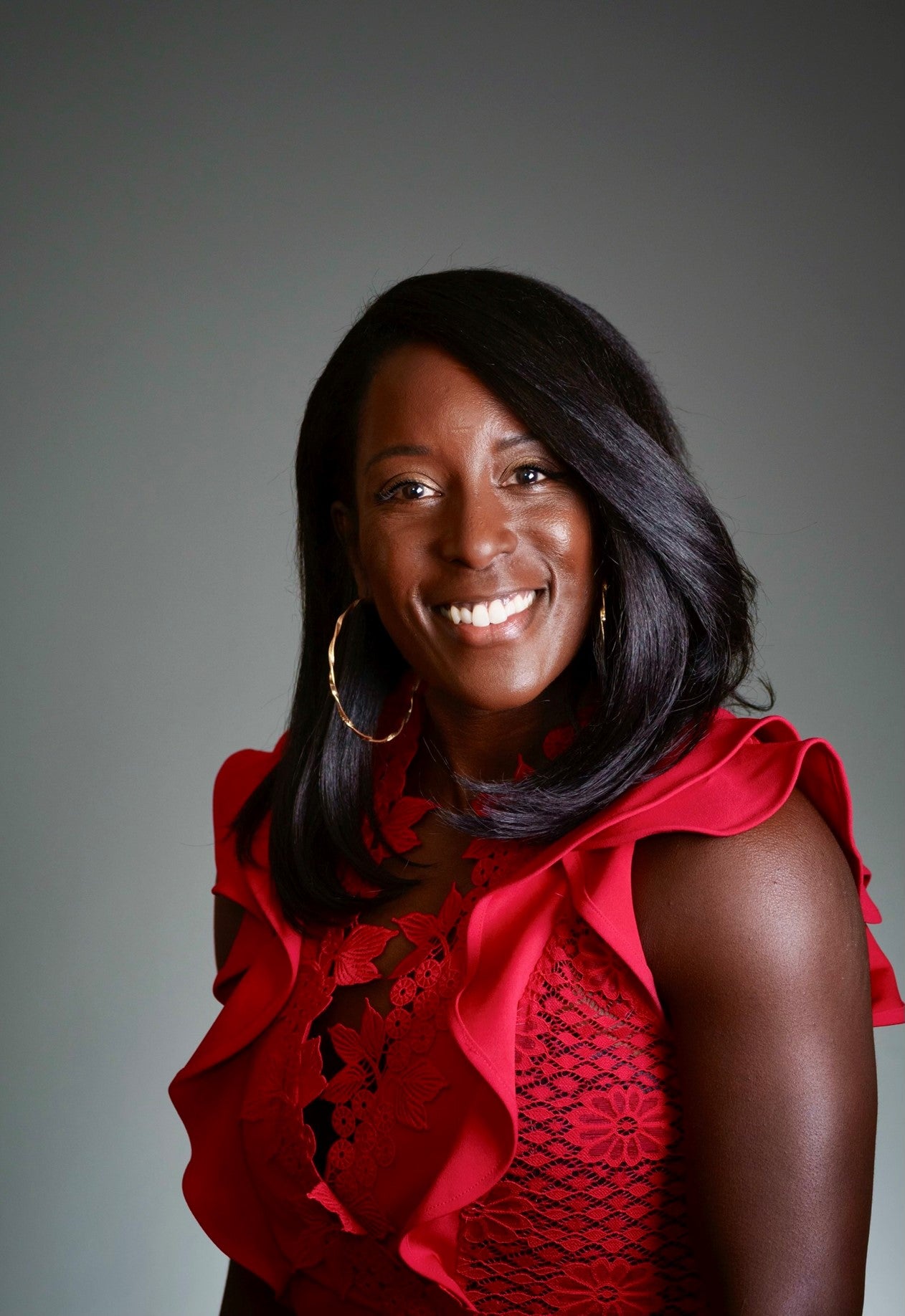 Tonnett Luedtke, a african american female-presenting individual, looks into the camera smiling. She is wearing a red pantsuit and has black hair.