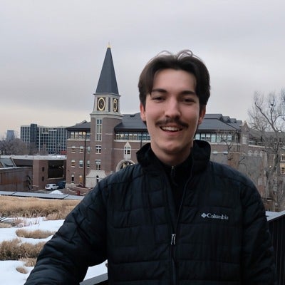 A male presenting individual standing in front of the law school building looking into the camera and smiling.
