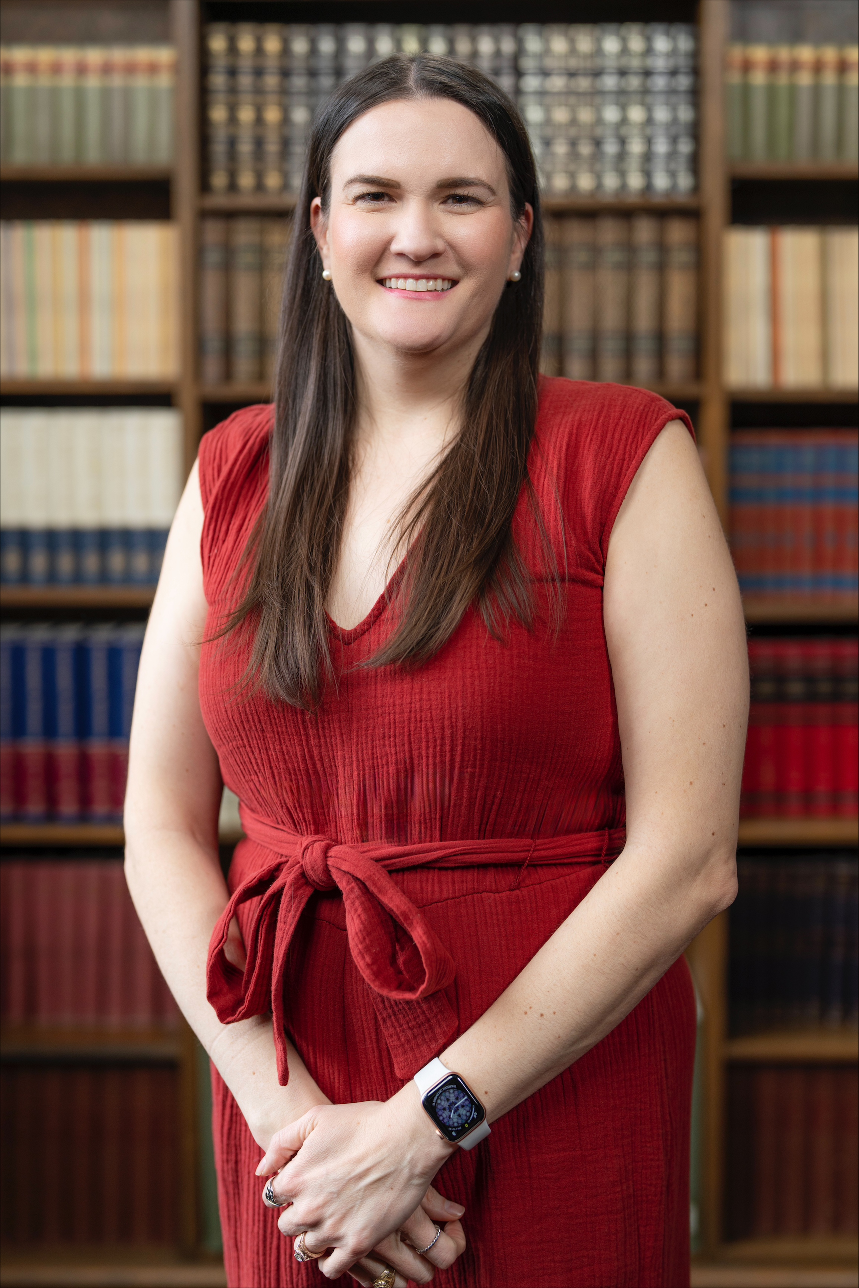 A female presenting individual, with brown hair and wearing a red jumpsuit, smiling into the camera. She is standing in front of a book case. 