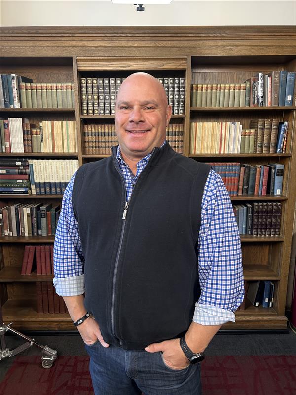 a male presenting individual standing in front of a bookcase smiling into the camera. wearing a black vest and blue shirt. 