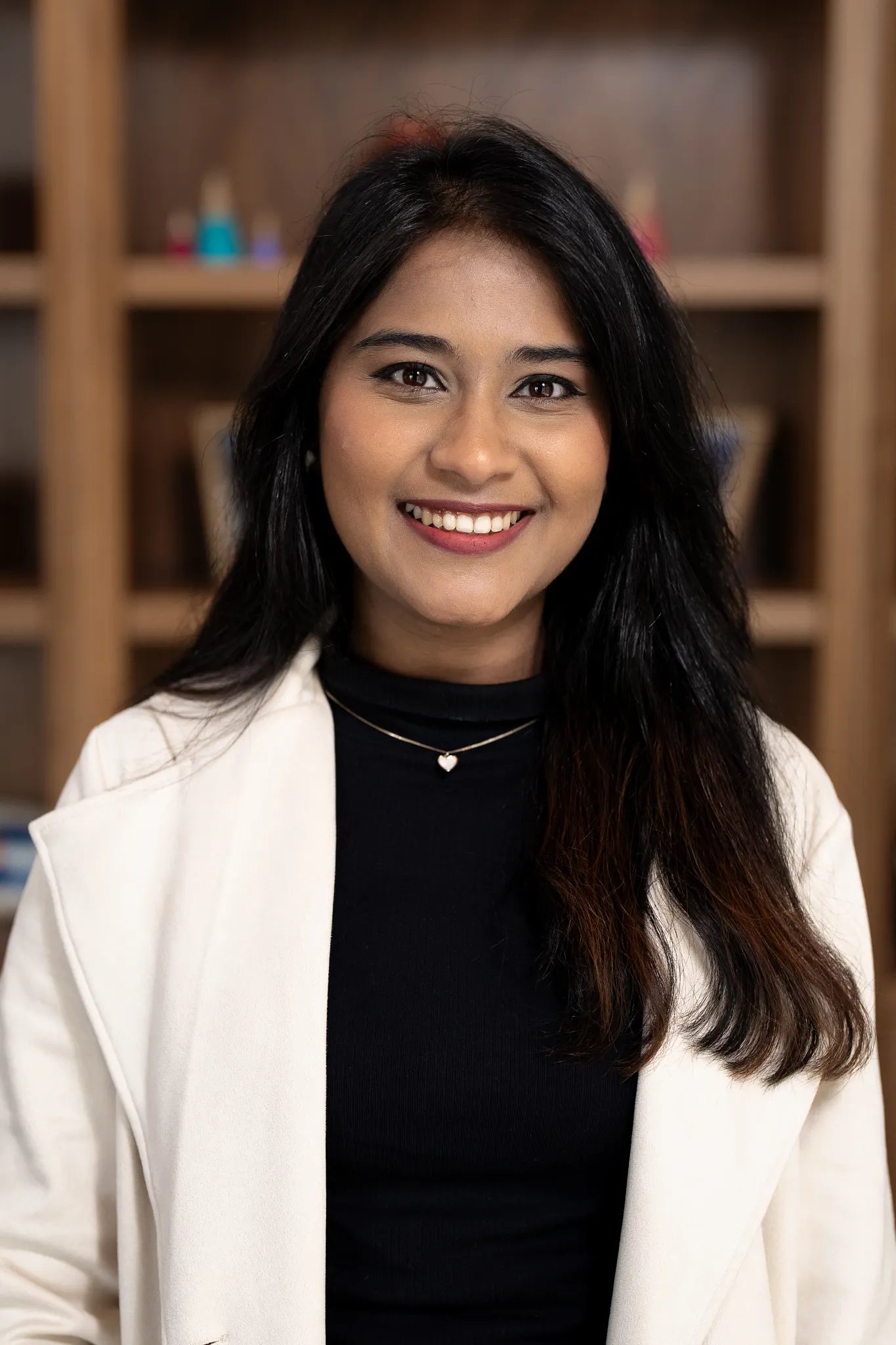 a female presenting individual with long black hair and brown eyes is smiling into the camera standing in front of a bookshelf. She is wearing a black blouse and white cardigan.