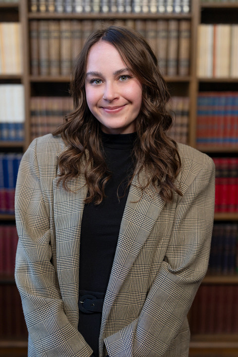 A female presenting individual with medium length wavy brown hair is smiling into the camera. She is wearing a tan blazer and black shirt. She is standing in front of a bookcase. 