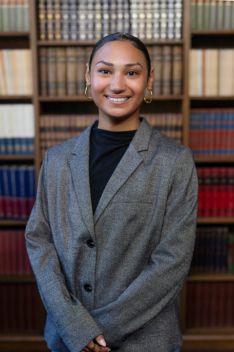 A female presenting woman wearing a grey jacket and black shirt looking into the camera smiling. She has black hair. She is standing in front of a bookcase.