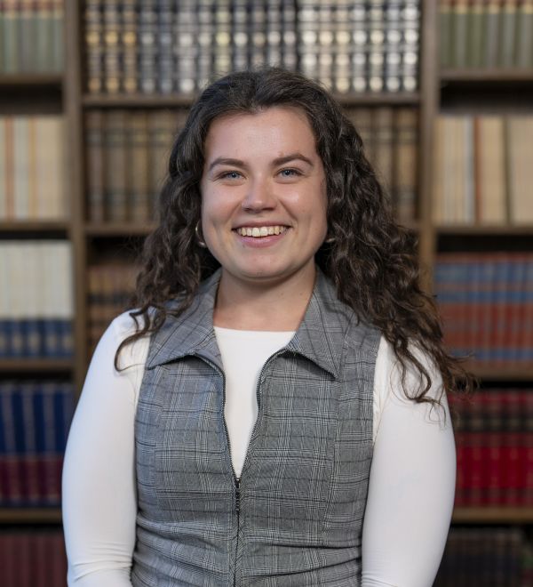 a white female presenting student with long brown curly hair is smiling into the camera. She is wearing a grey vest and is standing in front of a bookcase. 