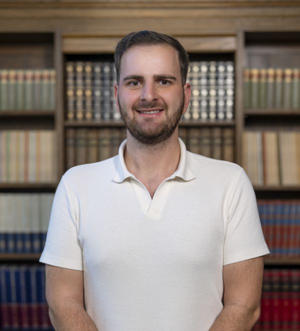 A white male presenting student wearing a white polo shirt is smiling into the camera standing in front of a bookcase.