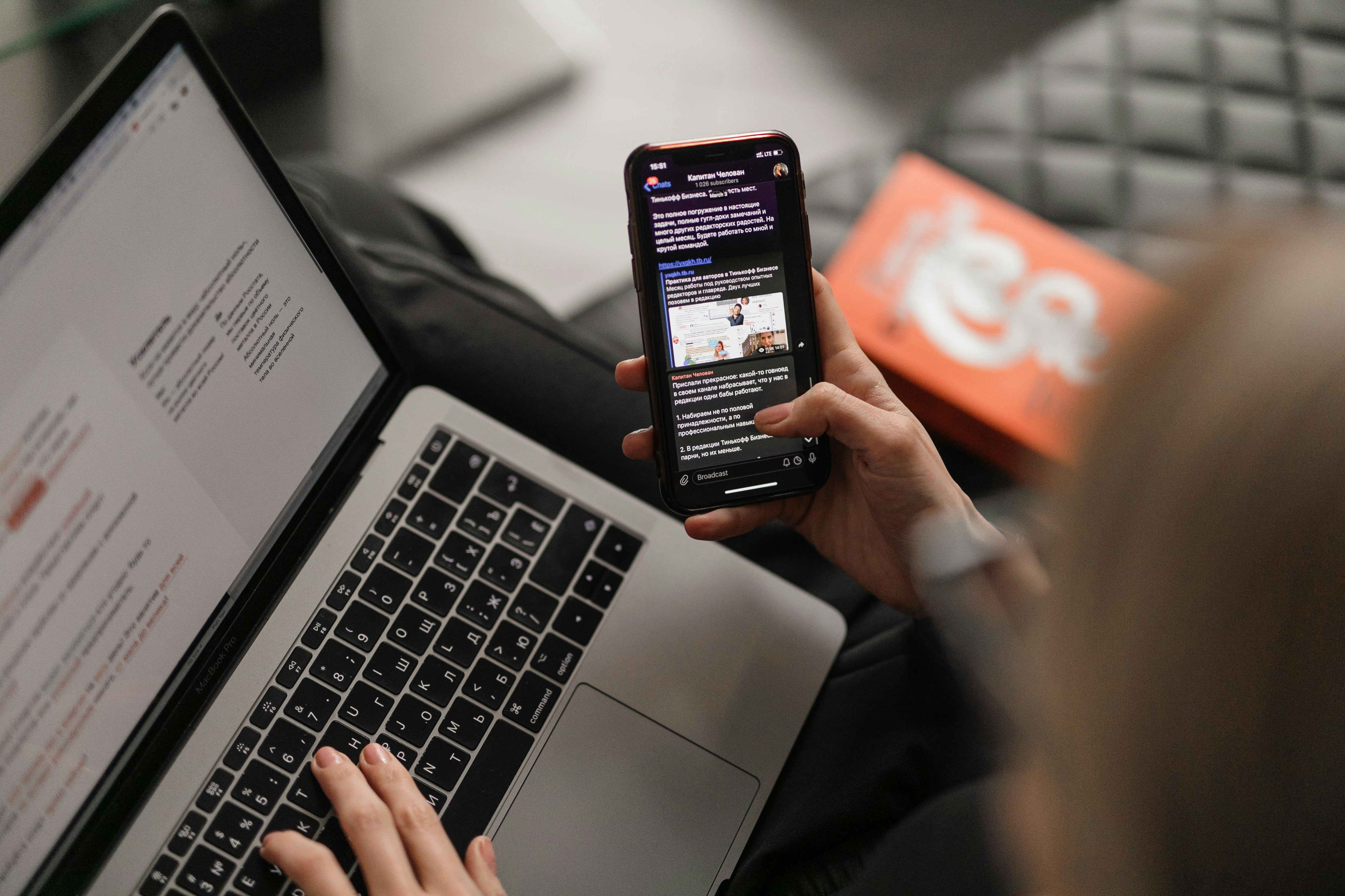 A person is sitting with a laptop on their lap, typing with one hand while holding a smartphone in the other. The laptop screen displays a document with visible text, and the phone screen shows a messaging app or social media post in a different language. The background is slightly blurred, and there is an orange box partially visible on the right side.