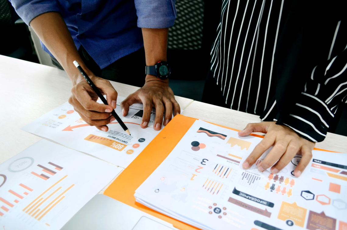 Close-up image of two individuals collaborating on a project. One person is pointing at a document containing charts, graphs, and data visualizations, while another person's hand is resting on the table. The setting suggests teamwork and engagement in a community or research initiative.