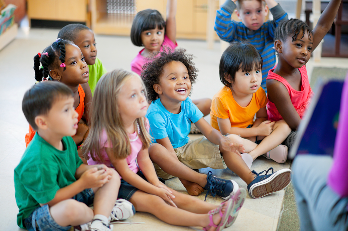 A group of diverse young children sitting together on the floor in a classroom, listening attentively to a teacher or storyteller who is partially visible in the foreground. The children display various expressions of curiosity, engagement, and interest. The classroom setting is bright and inviting.