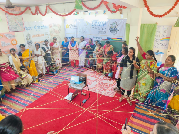 A group of women stands in a circle in a colorful, decorated room, holding interconnected strings to form a web-like structure. The floor is covered with vibrant striped mats, and a small table with a laptop and papers sits in the center of the circle. The background features banners with logos and text, as well as orange garlands hanging from the ceiling. The atmosphere suggests a community-building or teamwork activity.