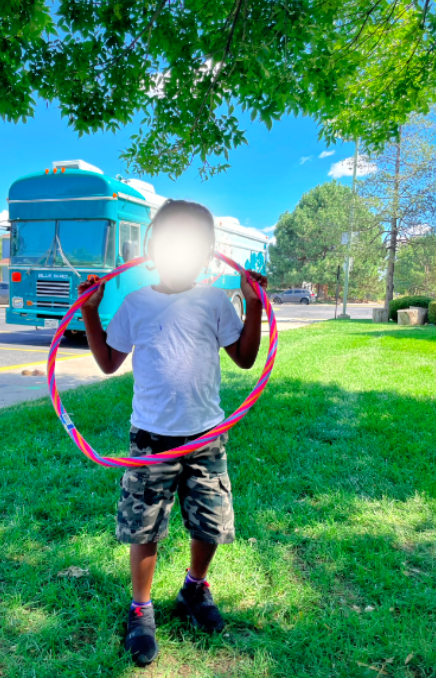 A young child is standing on grass under a tree, holding a pink hula hoop. The child is wearing a white t-shirt, camouflage shorts, and sneakers. A bright blue bus is parked in the background, and the sky is clear and sunny with trees and a street in the distance.