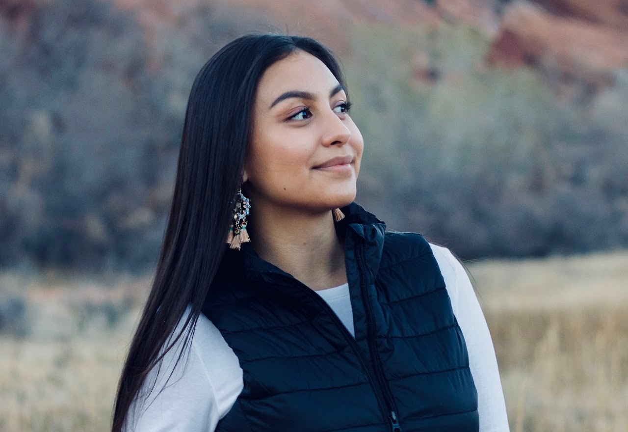 A woman with long, straight hair wearing a black puffy vest and a white shirt, looking to the side with a serene expression. She has decorative earrings and stands outdoors with a blurred natural background.