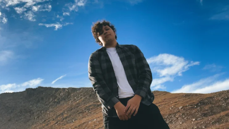 A young man is standing outdoors with a mountainous landscape in the background. He is wearing a plaid shirt over a white T-shirt, and his hands are casually clasped in front of him. The sky is clear with a few clouds, and the lighting suggests it's a bright day. The image is taken from a slightly low angle, making the man appear confident and relaxed.