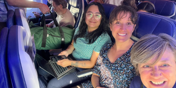 Three women on a plane, in their seats, smiling and taking a selfie.