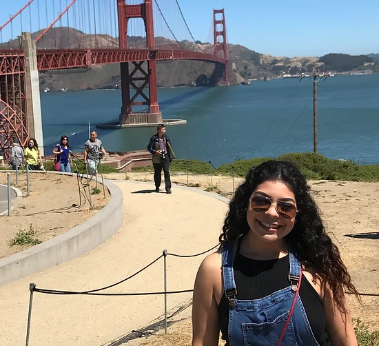 A woman with curly hair wearing sunglasses and denim overalls, smiling in front of the Golden Gate Bridge with a clear blue sky and water in the background. Other people are visible on the path behind her.