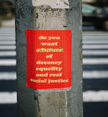 The image shows a red poster stuck on a street pole. The poster contains text in yellow and white that says, "Do you want decency, equality, and real social justice?" The background appears to be a street crossing with blurred lines and cars in the distance.