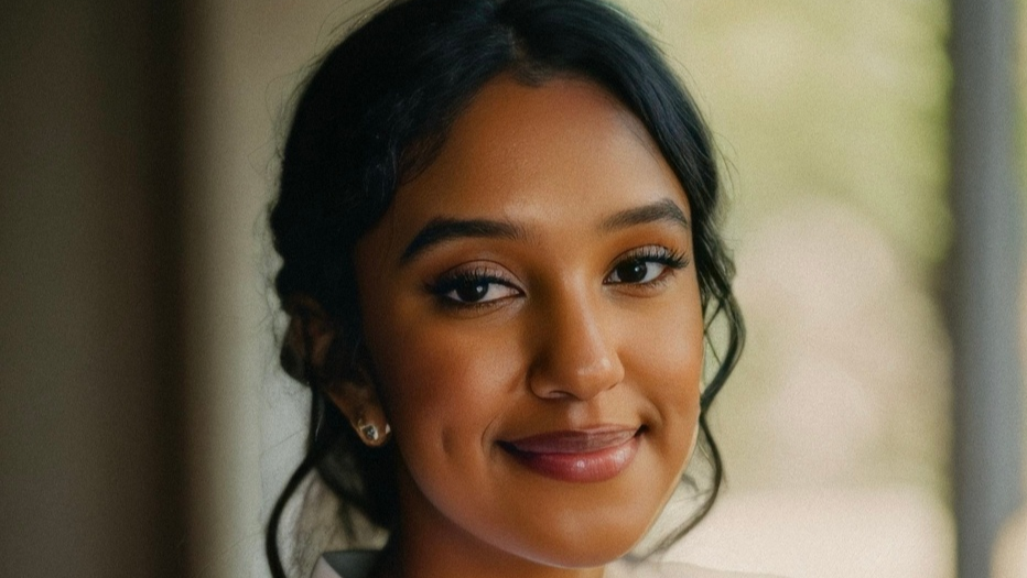 A close-up portrait of a young woman with a warm smile. She has dark, wavy hair pulled back and is wearing subtle makeup. The background is softly blurred, emphasizing her face. She is dressed in a light-colored top or jacket.