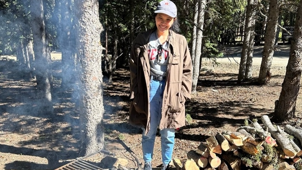 A person stands outdoors in a forested area next to a fire pit with a small fire burning. They are wearing a long brown coat, light blue jeans, and a gray cap, smiling at the camera. A stack of firewood is nearby, with trees and sunlight in the background. Smoke from the fire is visible in the air.