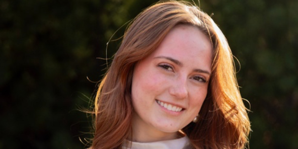 A young woman is dressed in a cream blouse posing with her arms at her side with her hands clasped together. She's standing in front of shrubbery looking and smiling directly at the camera.