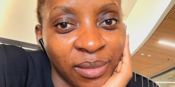 A young woman is taking a selfie, smiling slightly. She is dressed in a back blouse, with her left hand placed under her chin.