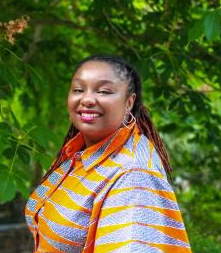 Black person outside smiling at camera with silver and orange striped top