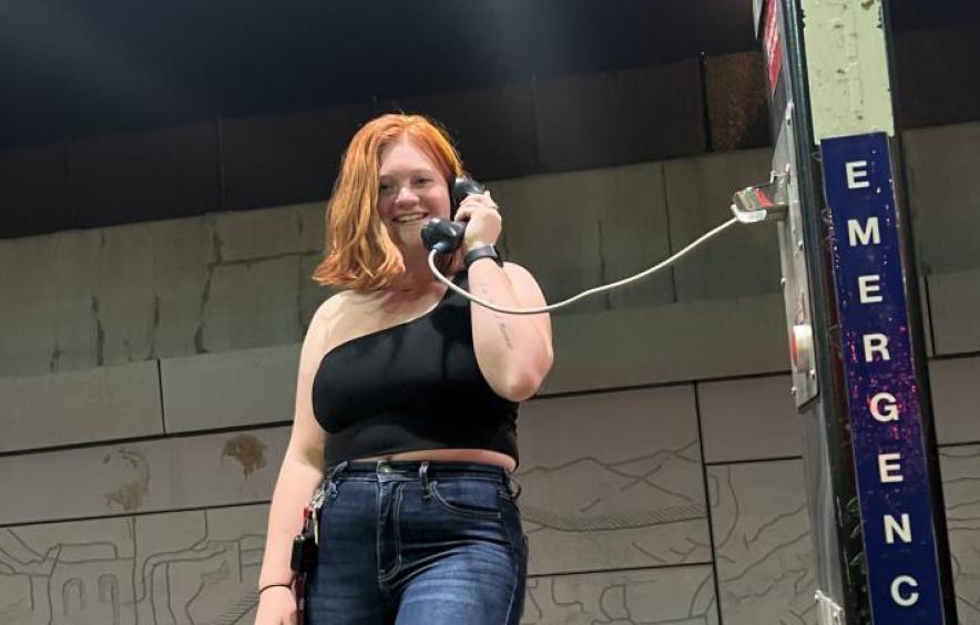 Allison Jones, a female-presenting person, smiles for the camera while holding her ear up to a public payphone