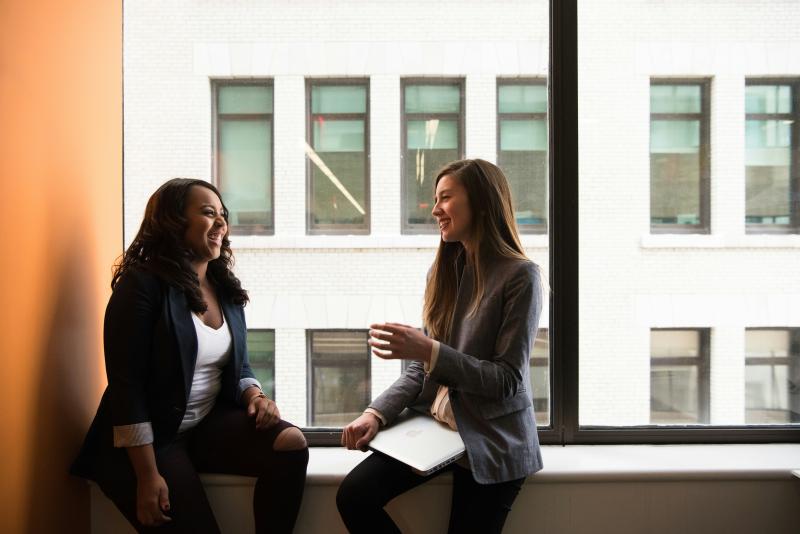 Two women sitting by a window sill wearing business attire. One woman is younger than the other, signaling a mentor and mentee relationship