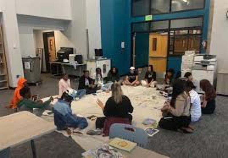 A group of students and a facilitator sit in a circle on the floor inside a school or library setting, engaged in a collaborative activity or discussion. Books and papers are spread out on a large cloth in the center, suggesting an interactive, inclusive, and reflective learning environment.