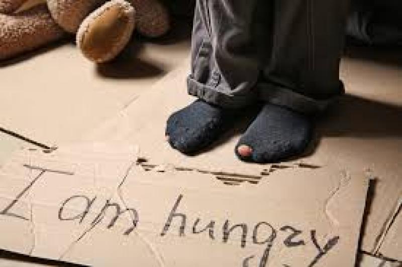 Child in torn socks standing on cardboard with the words "I am hungzy" written on it.