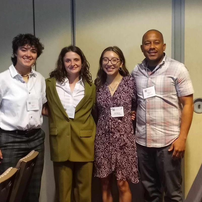 Four individuals stand closely together smiling for a photo in a professional setting. They are wearing name badges, suggesting attendance at a conference or formal event. The group consists of two women on the left, one woman in a floral dress and glasses, and a man in a plaid shirt on the right. The background shows a neutral-colored wall with a sliding door.