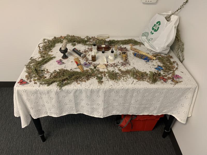 A lace-covered table with herbs, candles, bottles, decorative butterflies, and greenery; a recycling bag is visible to one side.