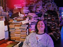 Zoe has shoulder-length, dark hair and is wearing a sweater over a graphic tee. She smiles in front of a tower of books that forms an arch.