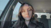 Bicky has long, dark hair and is sitting in a car.
