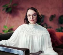 Jane has shoulder-length, dark hair and glasses and is wearing a white puffy shirt. She faces the camera in front of a mauve background.