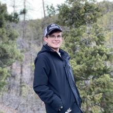 Man in glasses and hat stands in front of trees and smiles at the camera