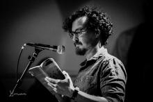 In a black and white image, a person with dark, curly hair, facial hair, and glasses reads from a book into a microphone.