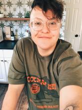 Tory has short, dark hair, round glasses, and a nose ring. They smile while wearing a green t shirt with orange lettering.