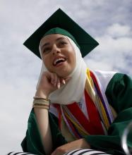 Fatma is wearing a white hijab and green graduation cap and gown. She rests her chin on her hand and smiles at something in the distance.