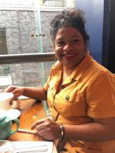 Renissa has dark hair pulled up in a bun and is wearing red lipstick and a mustard yellow collared top. She smiles while sitting at a table in a cafe.