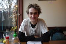 Monroe has tousled, short blond hair and light facial hair. He is wearing a black long-sleeve shirt under a white graphic tee, and he sits at a table in a cafe by a window and grins.