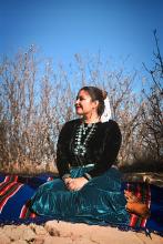 Char has her hair pulled up in a bun and sits on a colorful blanket outside. She is wearing a blue skirt, black top, and turquoise jewelry. 