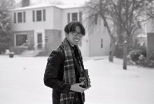 In this black and white image, Rishi stands smiling in front of a house that is out of focus in the background while it snows. He has short, dark hair, is wearing a winter coat and scarf, and holds a camera.