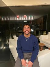 A person with short, curly hair and light facial hair wearing a blue sweater sits on the edge of a white couch and grins.