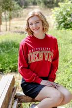 Kylie has short, curled, blond hair. She wears a red University of Denver sweatshirt and smiles as she sits on a bench.