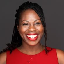 Mary has dark hair pulled back from her face. She is wearing a red top and red lipstick as she smiles brilliantly.