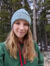 Sarah has shoulder-length, blond hair and is wearing a blue winter hat and green sweatshirt. She smiles softly while standing in a pine forest.